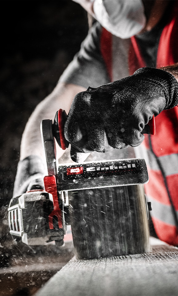 A person wearing black gloves sands a piece of wood using a red Einhell sander, with dust particles flying in the air.